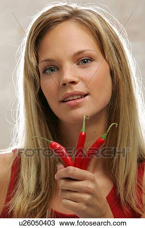 Stock Image - Young woman with long blond hair holding three chili peppers, portrait. Fotosearch