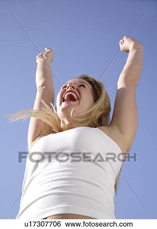 Young woman cheering with arms raised in front of blue sky, view from below View Large Photo Image Stock Photograph - Young woman cheering with arms raised in front of blue sky, view from below. Fotosearch