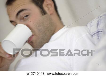 Stock Image - Young man sitting in bed, drinking mug of coffe, close up. Fotosearch