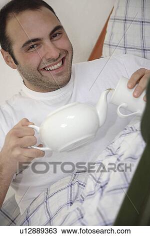 Young man sitting in bed, pouring cup of tea from teapot View Large Photo Image Stock Image - Young man sitting in bed, pouring cup of tea from teapot. Fotosearch