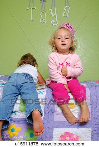 Stock Photo - Portrait of a girl sitting on the bed with her sister lying on the bed beside her. Fotosearch