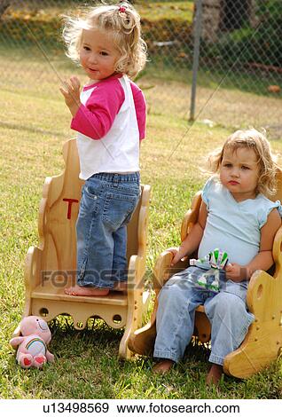 Stock Photo - Girl sitting in a chair with her sister standing on a chair beside her. Fotosearch
