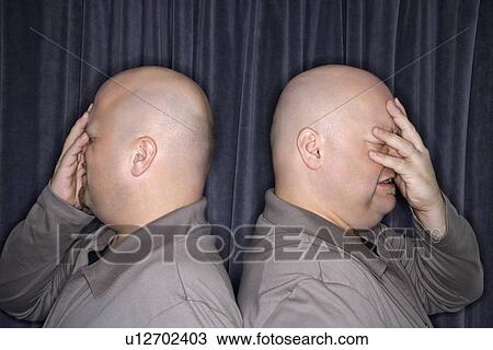 Stock Image - Profile of bald identical twin men standing back to back and grimacing with hands to head.. Fotosearch