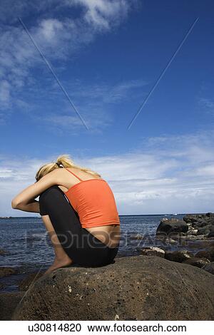 Stock Image - Caucasian young adult woman sitting hugging self on rocky shore.. Fotosearch