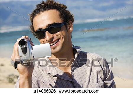 Stock Photograph - Mid-adult Caucasian man on beach pointing video camera at viewer.. Fotosearch