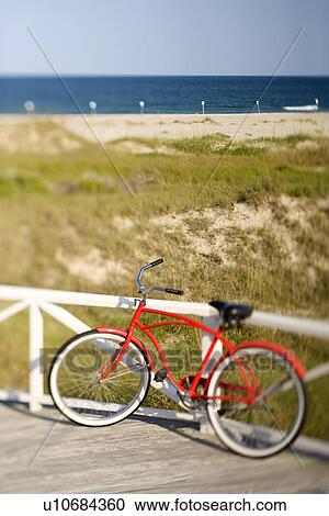 Stock Image - Red beach cruiser bicycle leaning against walkway rail on beach on Bald Head Island, North Carolina.. Fotosearch