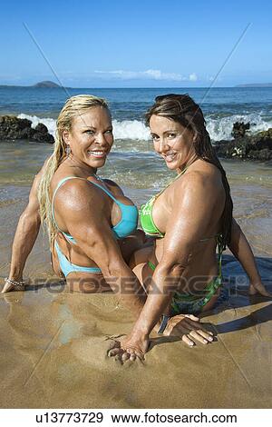 Stock Photo - Women bodybuilders in bikinis sitting in sand on beach. Fotosearch