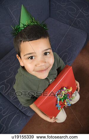 Stock Photo - Boy wearing party hat holding birthday present looking up . Fotosearch