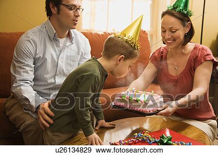 Stock Photo - Caucasian boy in party hat blowing out candles on birthday cake with family watching.. Fotosearch
