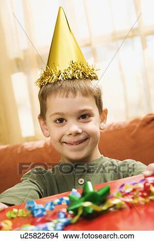 Picture - Caucasian boy wearing party hat holding gift and smiling at viewer.. Fotosearch