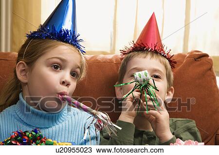 Stock Image - Caucasian children looking bored wearing party hats and blowing noisemakers.. Fotosearch