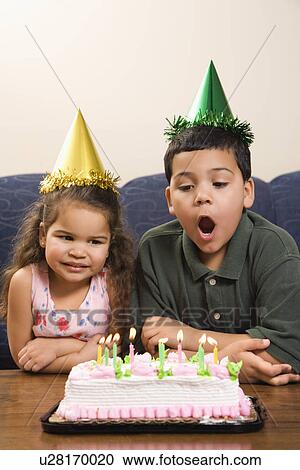 Stock Image - Girl and boy wearing party hats preparing to blow candles out on birthday cake. Fotosearch