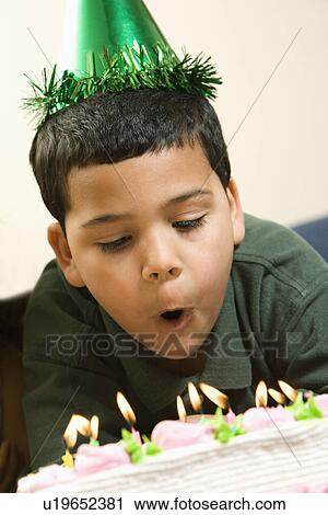 Stock Image - Hispanic boy wearing party hat blowing out candles on birthday cake.. Fotosearch