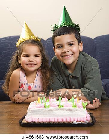 Stock Image - Hispanic girl and boy wearing party hats sitting in front of birthday cake smiling and looking at viewer.. Fotosearch