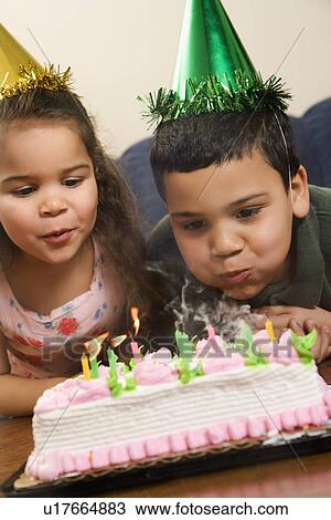 Stock Image - Hispanic girl and boy wearing party hats blowing out candles on birthday cake.. Fotosearch