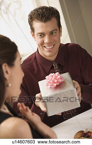 Stock Image - Mid adult Caucasian man presenting wrapped gift to surprised woman at restaurant.. Fotosearch