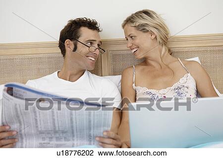 Stock Image - Mid adult couple using laptop and reading newspaper in bed. Fotosearch