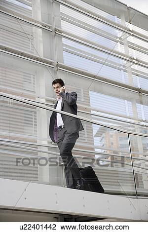 Stock Image - Businessman with suitcase talking on cell phone (low angle view). Fotosearch
