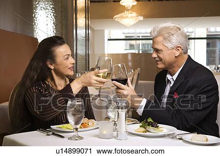 Stock Image - Caucasian mature adult male and prime adult female sitting at restaurant table toasting wine glasses.. Fotosearch