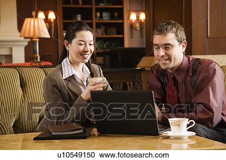 Stock Image - Caucasian mid adult businessman and woman drinking coffee and looking at laptop computer.. Fotosearch