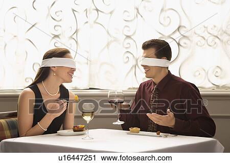 Stock Image - Mid adult couple dining in a restaurant with blindfolds over eyes. Fotosearch