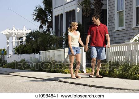 Stock Image - Mid-adult couple walking on suburban sidewalk holding hands. Fotosearch