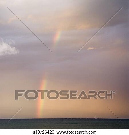 Stock Photograph - Rainbow spreading across sky on coast. Fotosearch