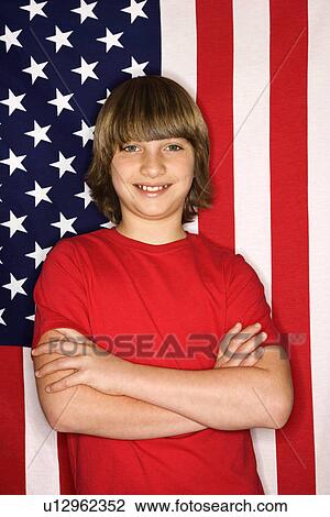 Stock Image - Portait of Caucasian boy with arms crossed with American flag in background.. Fotosearch