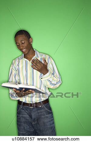 Stock Photo - Portrait of African-American teen boy reading book standing in front of green background.. Fotosearch