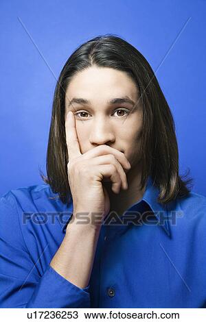 Stock Image - Portrait of teen boy with hand covering chin and finger on side of face against blue background. Fotosearch