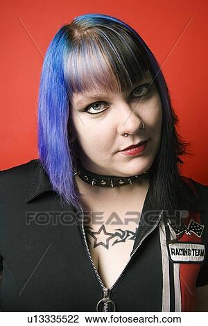 Stock Image - Portrait of woman with blue hair, tattoo, and spike collar against red background. Fotosearch