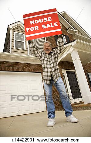 Stock Image - Male outside house holding up for sale sign. Fotosearch