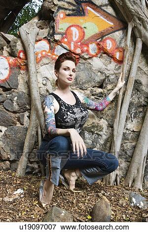 Sexy tattooed woman crouching next to wall covered in graffiti and Banyan tree branches View Large Photo Image Stock Photo - Sexy tattooed woman crouching next to wall covered in graffiti and Banyan tree branches. Fotosearch