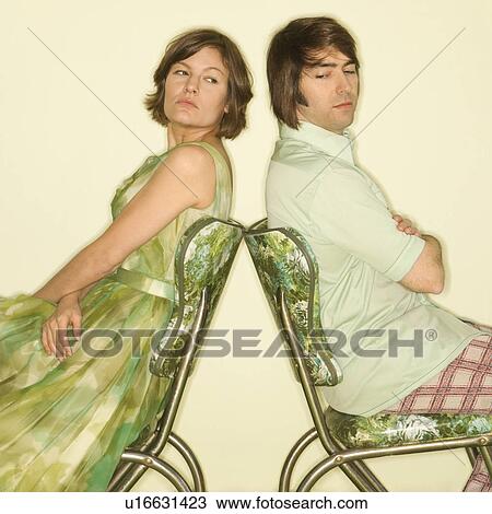 Stock Image - Caucasian mid-adult couple wearing vintage clothing sitting back to back in green vinyl chairs with arms crossed looking angry.. Fotosearch