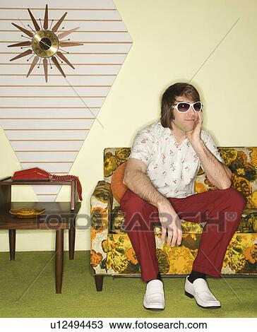 Stock Image - Caucasian mid-adult man wearing sunglasses sitting on colorful retro sofa looking bored.. Fotosearch