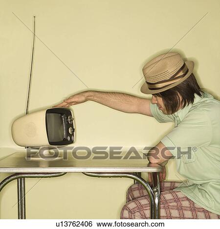 Stock Photograph - Man wearing hat sitting at 50's retro dinette set tapping old television set. Fotosearch