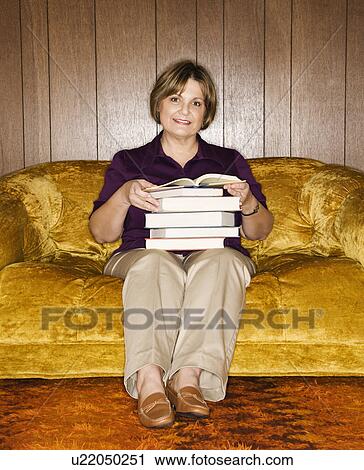 Stock Image - Woman sitting on couch in living room holding stack of books.. Fotosearch