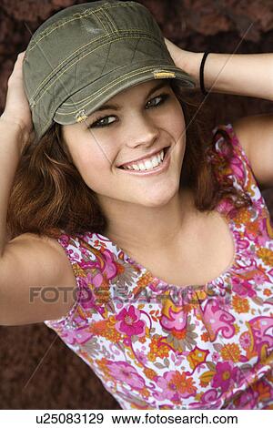 Stock Photo - Young Caucasian woman wearing hat with hands behind head smiling at viewer.. Fotosearch