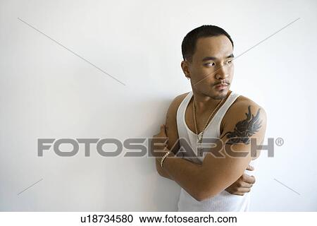 Stock Image - Portrait of young adult man leaning against white wall with arms crossed looking over shoulder. Fotosearch