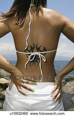Stock Photography - Rear view of multi ethnic young adult woman on beach with hands on hips and tattoo.. Fotosearch