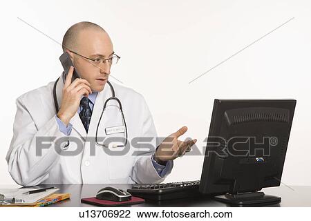 Stock Image - Caucasian mid adult male physician sitting at desk with laptop computer talking on cellphone.. Fotosearch