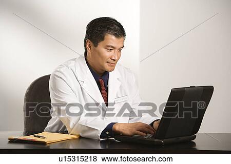 Stock Photo - Man in a lab coat typing on a laptop computer at a desk. Fotosearch