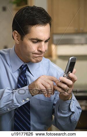 Stock Photograph - MId-adult Caucasian male pressing cell phone buttons while standing in kitchen.. Fotosearch
