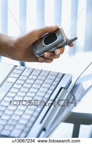 Picture - Close up of young woman's hand holding cellphone with laptop in background. Fotosearch