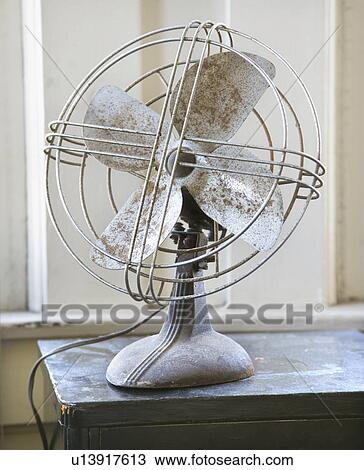 Stock Image - Retro style metal fan sitting on tabletop by window.. Fotosearch