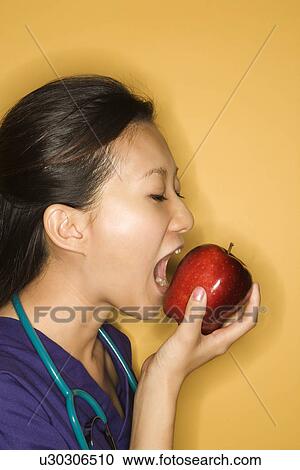 Stock Image - Asian Chinese mid-adult female doctor taking bite of red apple against yellow background.. Fotosearch