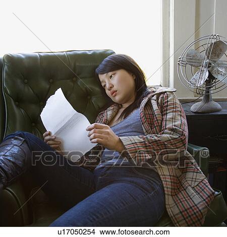 Picture - Pretty young Asian woman sitting in green chair reading paper.. Fotosearch