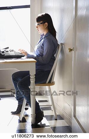 Stock Image - Pretty young Asian woman sitting in chair typing on typewriter in kitchen.. Fotosearch