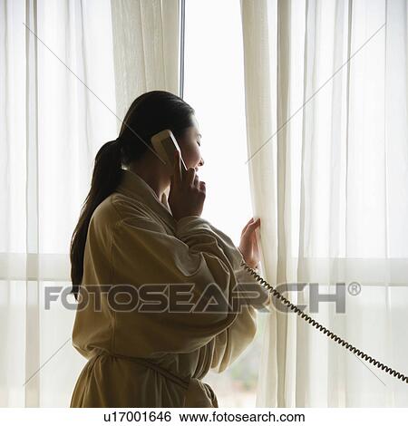 Stock Photograph - Woman in bathrobe talking on phone and looking out window. Fotosearch