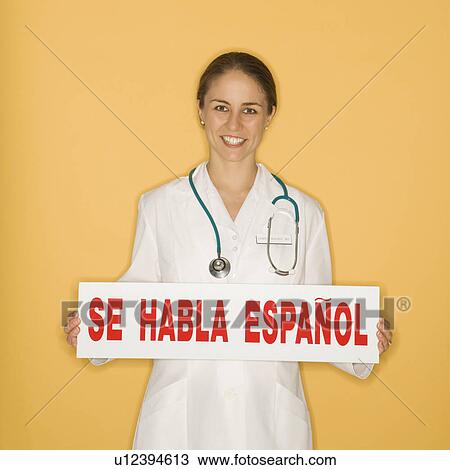 Stock Image - Portrait of doctor holding up se habla espanol sign against yellow background. Fotosearch